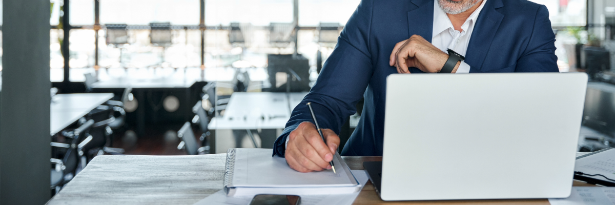 Man looking at a screen, writing on notepad. 