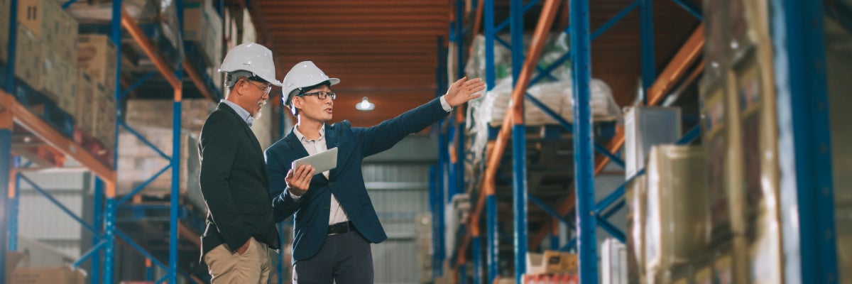 Two people standing in a warehouse with hardhats on. One person is holding an ipad, pointing to something in the distance.