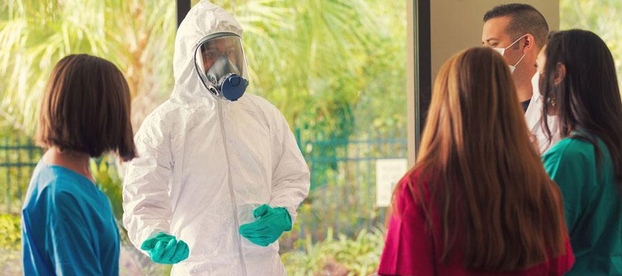 Communicable Disease Outbreak Prevention. A man in a full hazmat suit speaks to clinicians wearing surgical masks.